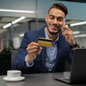 Happy young businessman holding credit card and talking on cellphone